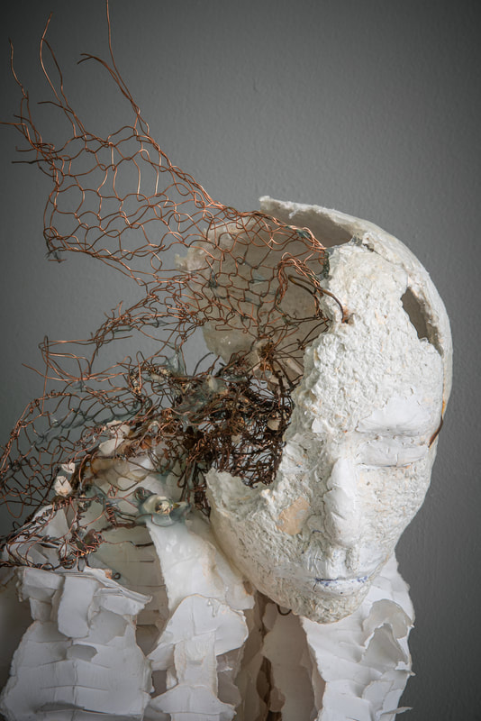 Head detail of a white sculpture with an amorphous neck, shoulders, and torso. One side of the head is broken open, and copper mesh wire fills the head cavity and extends in sharp points above the head.