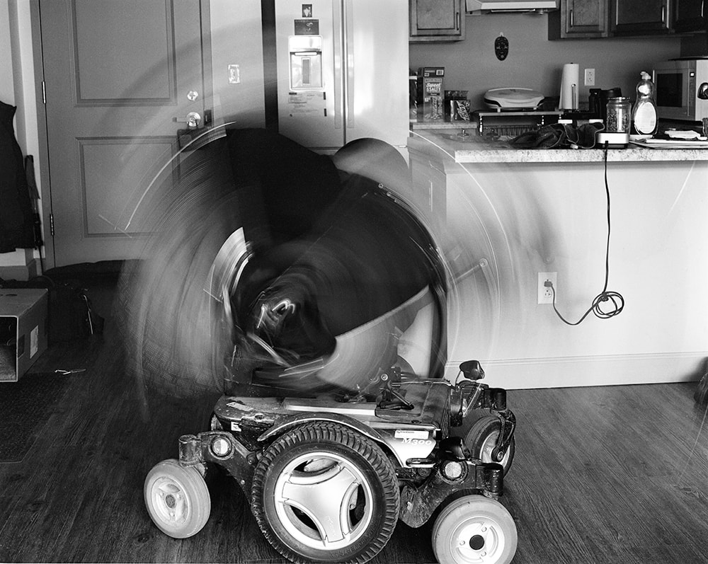 A black and white photo of a power wheelchair. The seat is blurred with the motion of rocking back and forth.