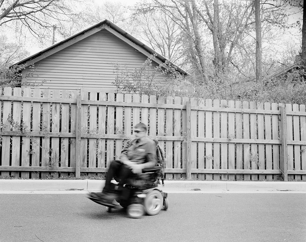 A black and white photo of a man in a power wheelchair riding in front of a wood fence. The man and his chair are blurred with forward motion.