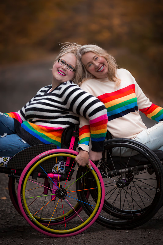 Two smiling pale skinned blonde women wearing striped sweaters sit in wheelchairs and lean their backs against each other.