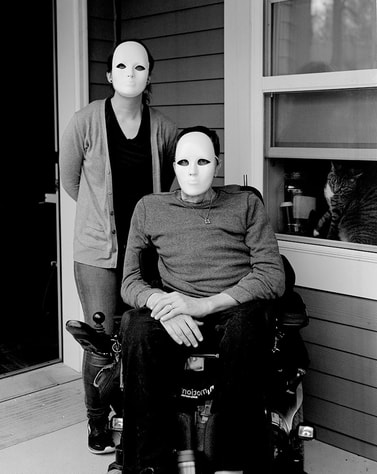 A black and white photo of two people in white masks. One stands at the left, the other sits in a wheelchair on the right.