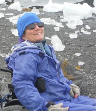 Photo of a smiling white woman wearing a blue cap and coat and a pair of sunglasses. She is in a wheelchair in front of an icy field.