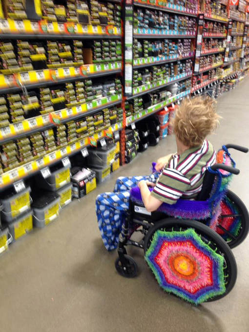 The bright yarn-bombed wheelchair with a blonde, tousled hair rider in front of a row of hardware shelves.
