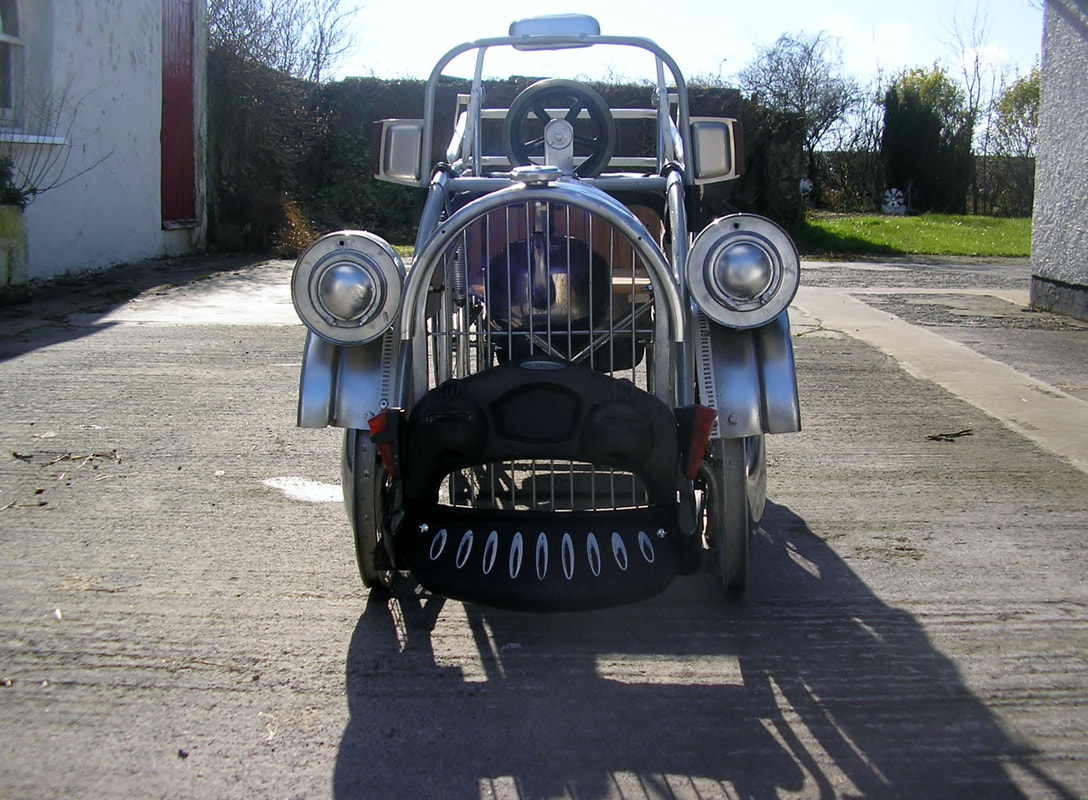 Back view of a wheelchair with additions to create the look of an antique car. It has 4 aluminum wheels, headlights, front grid and bumpers, and an open top with a steering wheel.
