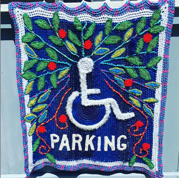 A crocheted blue blanket with a white wheelchair symbol in the middle surrounded by green leaves and red flowers.