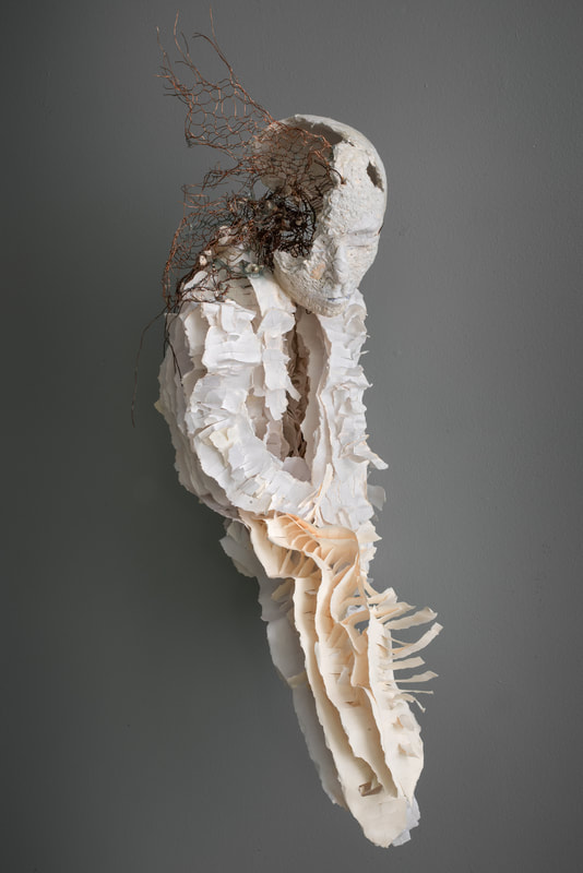 A white sculpture of a head with an amorphous neck, shoulders, and torso. The body is built of scraps of paper and resembles a wasps' nest. One side of the head is broken open, and copper mesh wire fills the head cavity and extends in sharp points above the head.