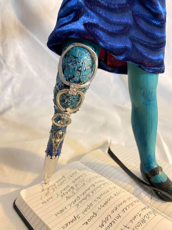 Leg detail of an art doll with a jeweled pen as a lower right leg. The doll wears aqua tights, a blue velvet skirt, and one black Mary Jane shoe on the left foot. The right leg/pen is writing in a small notebook.