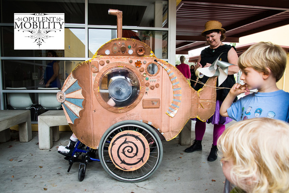 On the right, two blonde children look at a bronze submarine wheelchair costume. 