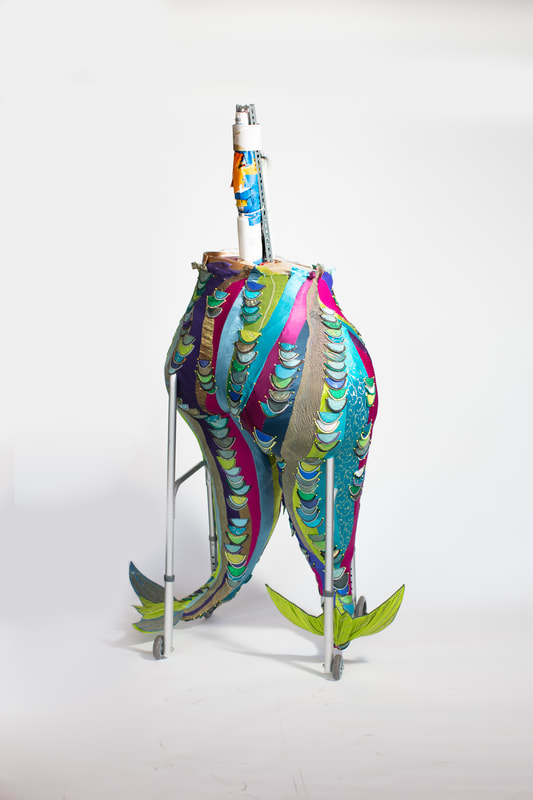Back view of the lower body of a figure with two legs/tail fins built into a wheeled walker. The figure is covered in colorful swirls of fabric with contrasting scales edged in zipper teeth.