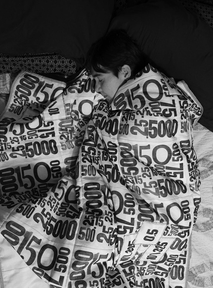 Black and white photo of a pale skinned person with dark hair lying on a bed, wrapped in a black and white quilt printed with numbers representing medication doses.