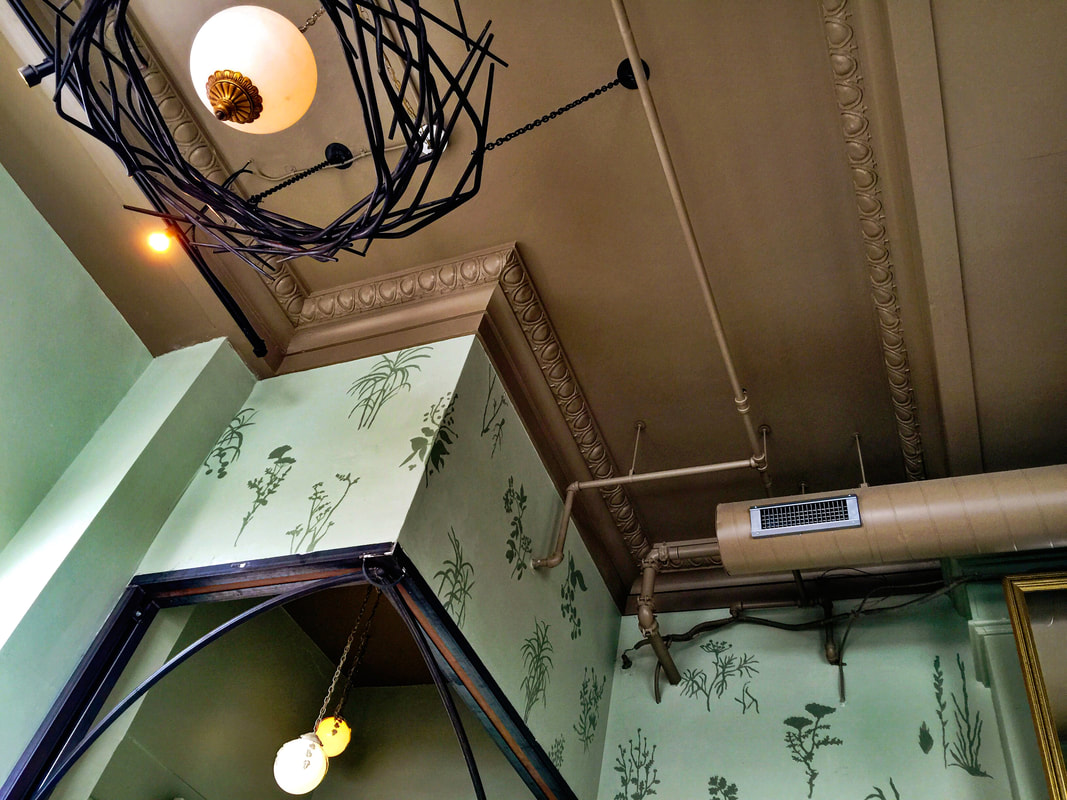 Looking up at a coppery brown ceiling with crown molding, a branch like light fixture, and pale green walls with darker green herbs and plants.