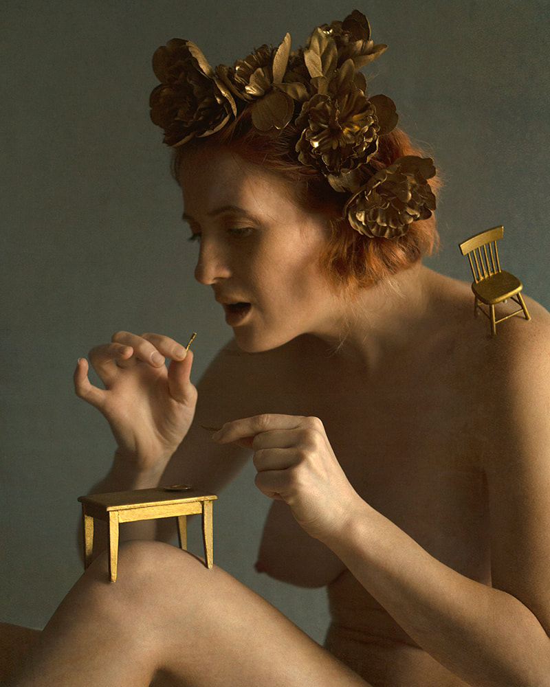 A nude, pale-skinned woman with a bronze floral headpiece. She is seated with a small golden table on one knee and attempts to eat from a tiny golden plate with a tiny fork and knife. On her shoulder is perched a small gold chair.