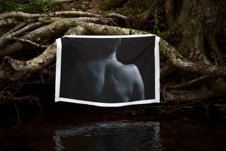A photo of a woman's back and shoulders hangs from knotted tree roots over water.
