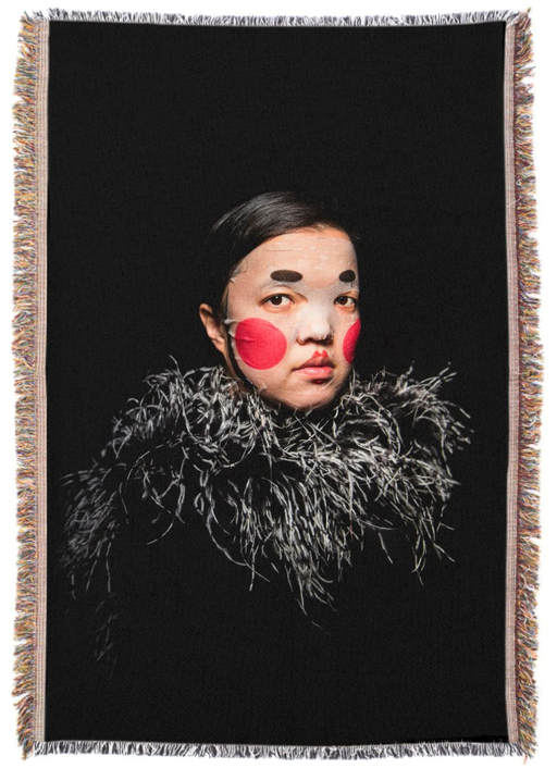 Black tapestry with a dark haired woman in a painted face mask with a grey feather boa around her neck.
