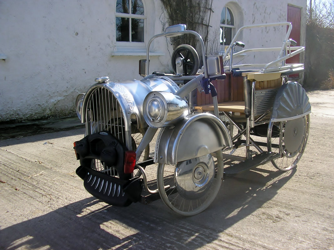Left side view of a wheelchair with additions to create the look of an antique car. It has 4 aluminum wheels, headlights, front grid and bumpers, and an open top with a steering wheel.