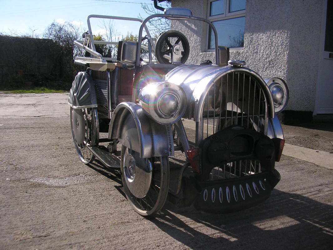 Right side view of a wheelchair with additions to create the look of an antique car. It has 4 aluminum wheels, headlights, front grid and bumpers, and an open top with a steering wheel.