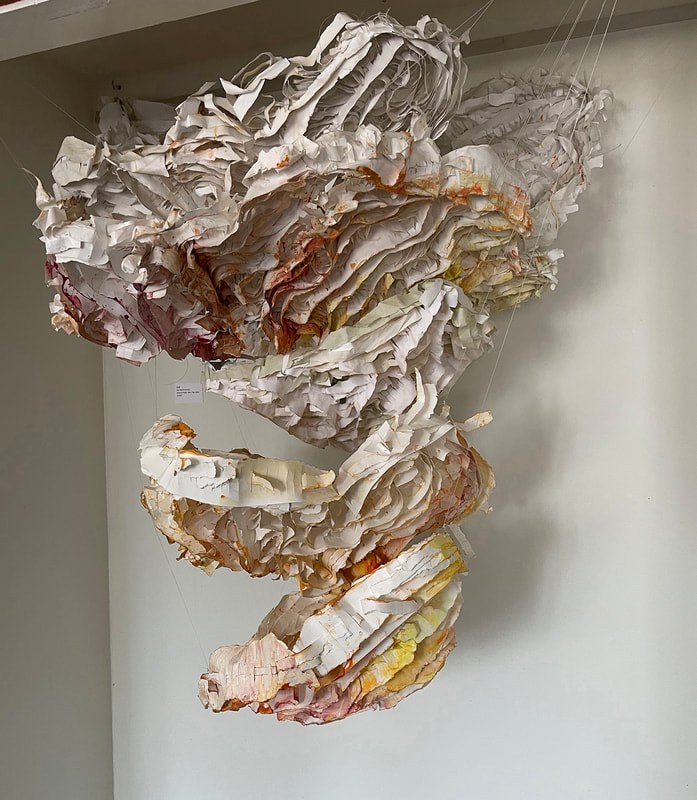 A bulbous spiral sculpture made of woven and tinted paper in white, tan, gold, and red.