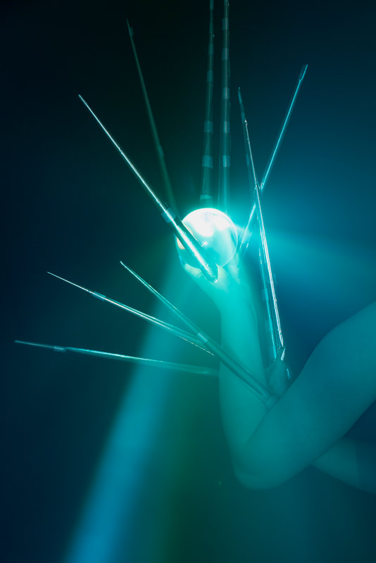 Pale arms and hands with long metallic pointed finger extensions hold a cluear orb up to a bright blue-green light.