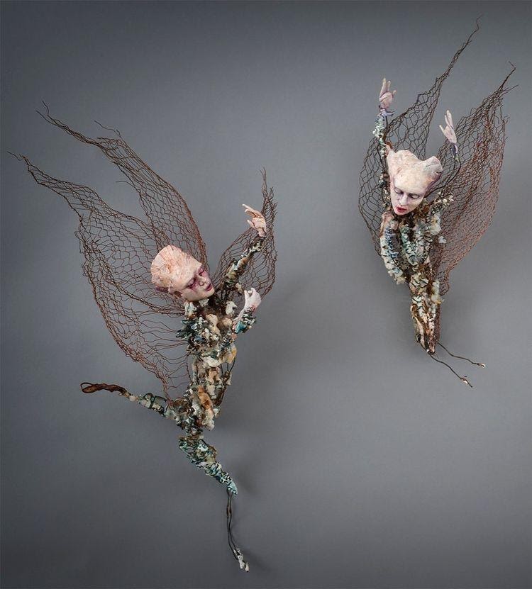Two figures with oversized painted heads, twisted bodies, and large copper wire wings against a grey background.