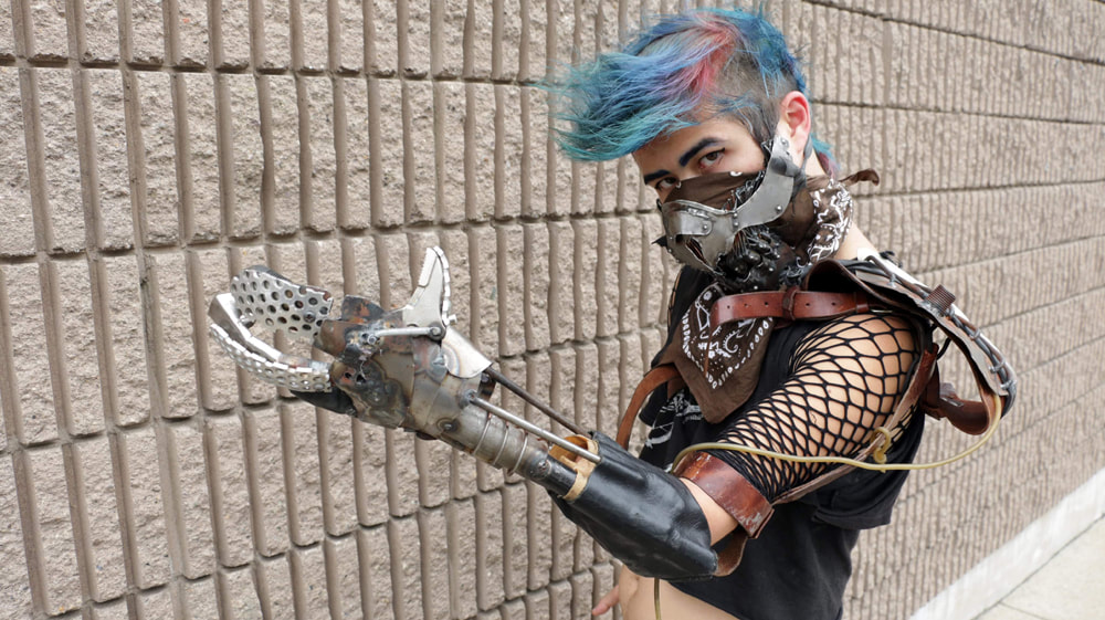 Side view of a white woman with blue and purple hair, a rough looking metal mask, and a rough metal and grid-work prosthetic arm against a concrete brick wall.