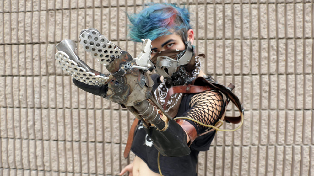 A white woman with blue and purple hair, a rough looking metal mask, and a rough metal and grid-work prosthetic arm against a concrete brick wall.