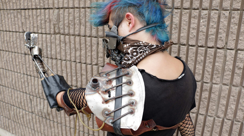 Back view of a white woman with blue and purple hair, a rough looking metal mask, and a rough metal and grid-work prosthetic arm against a concrete brick wall.
