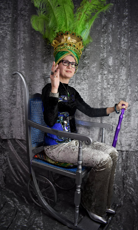 A person wearing a large green and gold feathered headdress sits in a silver wheelchair against a silvery grey backdrop. She holds a purple cane in her left hand.