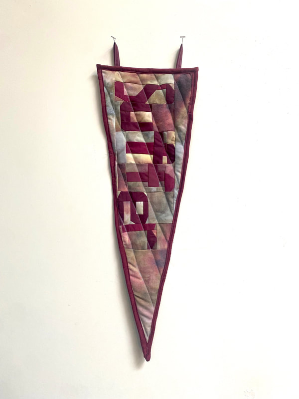 A burgundy, pink, yellow, and grey quilted triangular pennant with the word Suffer quilted down the center.