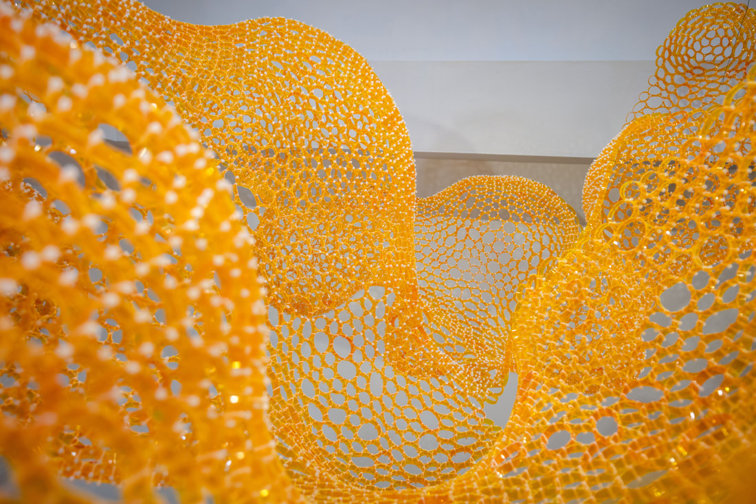 Undulating netting sculpture made of slices of yellow/orange pill bottles zip tied together.