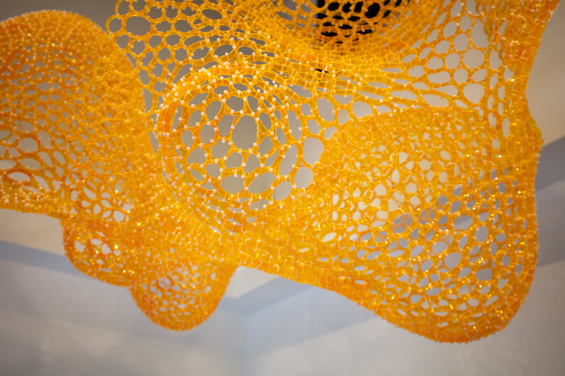 Second view of an undulating netting sculpture made of slices of yellow/orange pill bottles zip tied together.