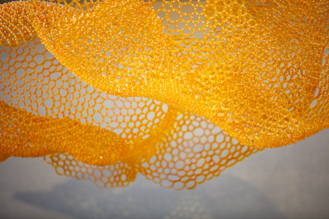 Underneath view of an undulating netting sculpture made of slices of yellow/orange pill bottles zip tied together.