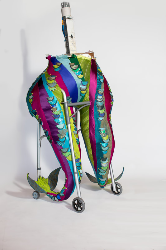 The lower body of a figure with two legs/tail fins built into a wheeled walker. The figure is covered in colorful swirls of fabric with contrasting scales edged in zipper teeth.