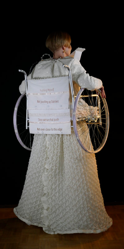 Back view of a pale skinned woman in a long white gown with a wheelchair attached to her waist and arms. The back of the wheelchair has lines of text printed on them: Pushing Myself, Not Pushing up Daisies, Sttop patriarchal push, Not even close ot the edge.