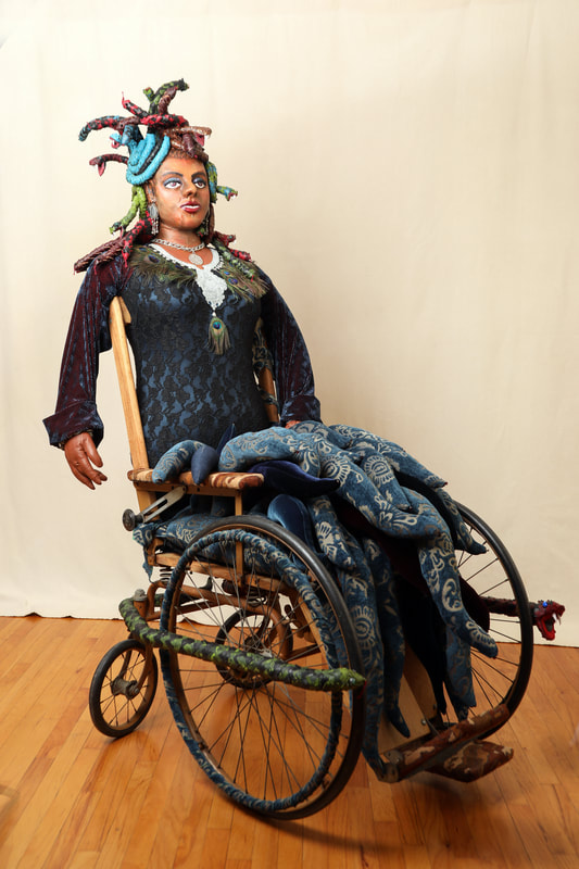 A sculpture of Medusa is built into the seat of a vintage wheelchair. The figure has snakes for hair, a peacock feather necklace, and a seat full of wave-like tentacles.