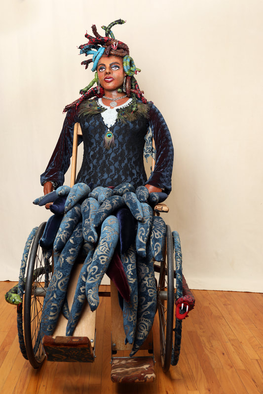 A sculpture of Medusa is built into the seat of a vintage wheelchair. The figure has snakes for hair, a peacock feather necklace, and a seat full of wave-like tentacles.