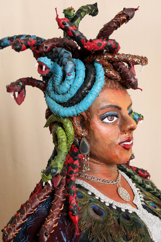 The head and shoulders of a Medusa sculpture, focused on the multi-colored leather snake hair, painted face, and silver and peacock feather necklace.