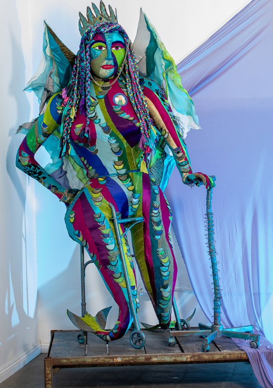 Sculpture of a multicolored winged woman with fishtail legs built into a walker and using a wheeled cane.