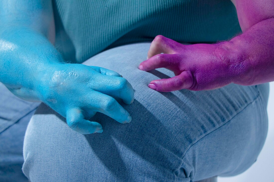 Two hands and arms with twisted fingers rest on top of legs wearing faded denim jeans. One arm and hand is sky blue and the other is a deep magenta.