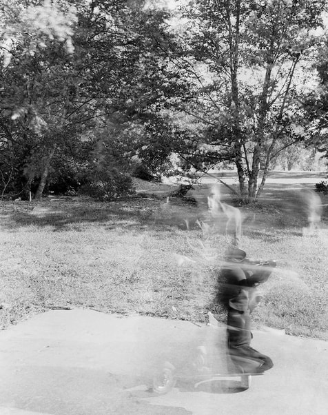 A black and white ghostly image of a man in a power wheelchair. His head and torso are faintly visible as swirls, but his left lower leg and foot are clear. In the background are grasses and trees.