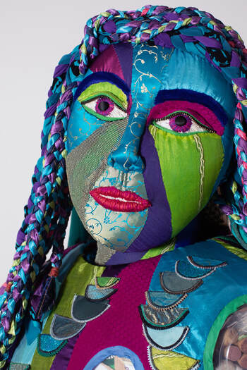 Bust of a sculpted female figure covered in swirls of green, blue, purple, magenta, and gold fabrics. Her purple eyes and red lips are embroidered, and her multi-colored hair is braided.