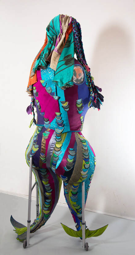 Back view of a sculpted female figure with two legs/tail fins built into a wheeled walker. The figure is covered in colorful swirls of fabric with contrasting scales edged in zipper teeth.