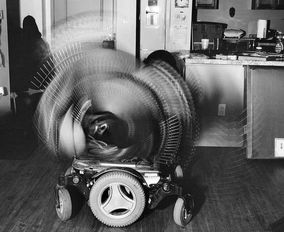 A black and white image of a power wheelchair on a hardwood floor. The top part of the wheelchair blurs as it tilts back and forth. In the background is a dark figure on the left and a kitchen counter on the right.