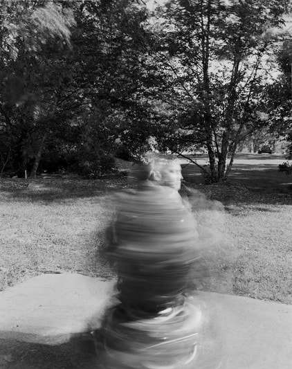 A black and white photo of a person spinning on a power wheelchair. The image is blurred, like the spinning of a tornado. In the background is grass and trees.