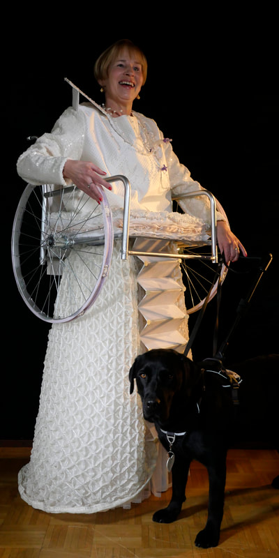 A pale-skinned woman in a long white dress with a wheelchair attached around her waist and arms. She holds the leash of a black guide dog.