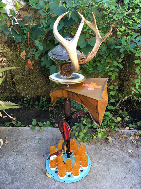A turtle shell and antlers mounted over an antique medicine chute on top of a hand cranked drill. The base is a pal blue disc covered with upside down pill bottles.