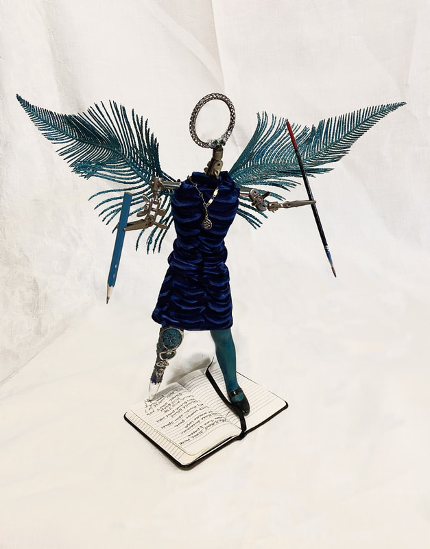 An art doll with turqouise wings, a soldering clamp stand for a head and arms, and a jeweled pen as a lower right leg. The doll wears aqua tights, a blue velvet dress, and one black Mary Jane shoe on the left foot. The right leg/pen is writing in a small notebook.